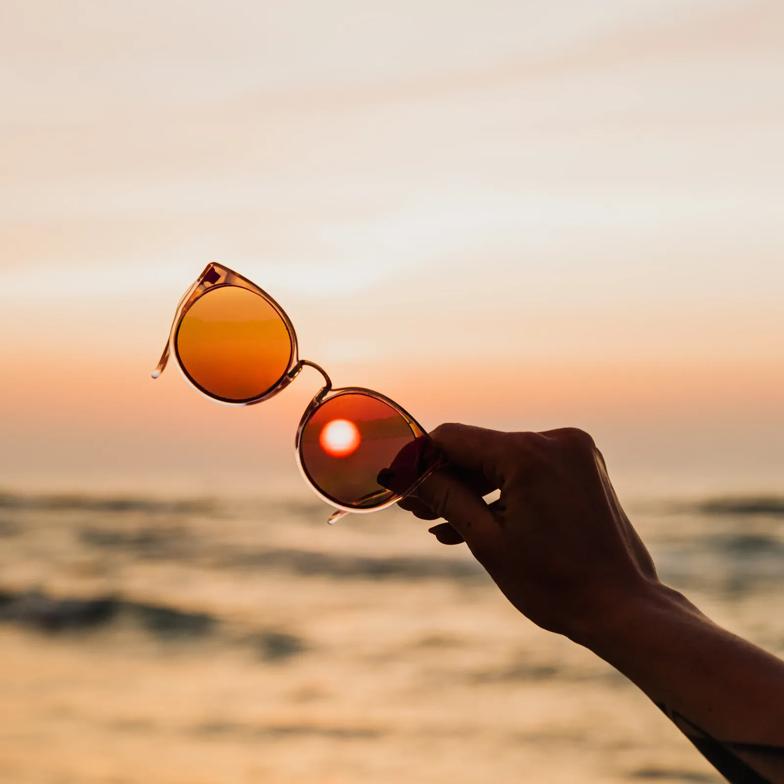 Mano que sostiene unas gafas de sol frente al mar al atardecer, con el sol visto a través de una de las lentes